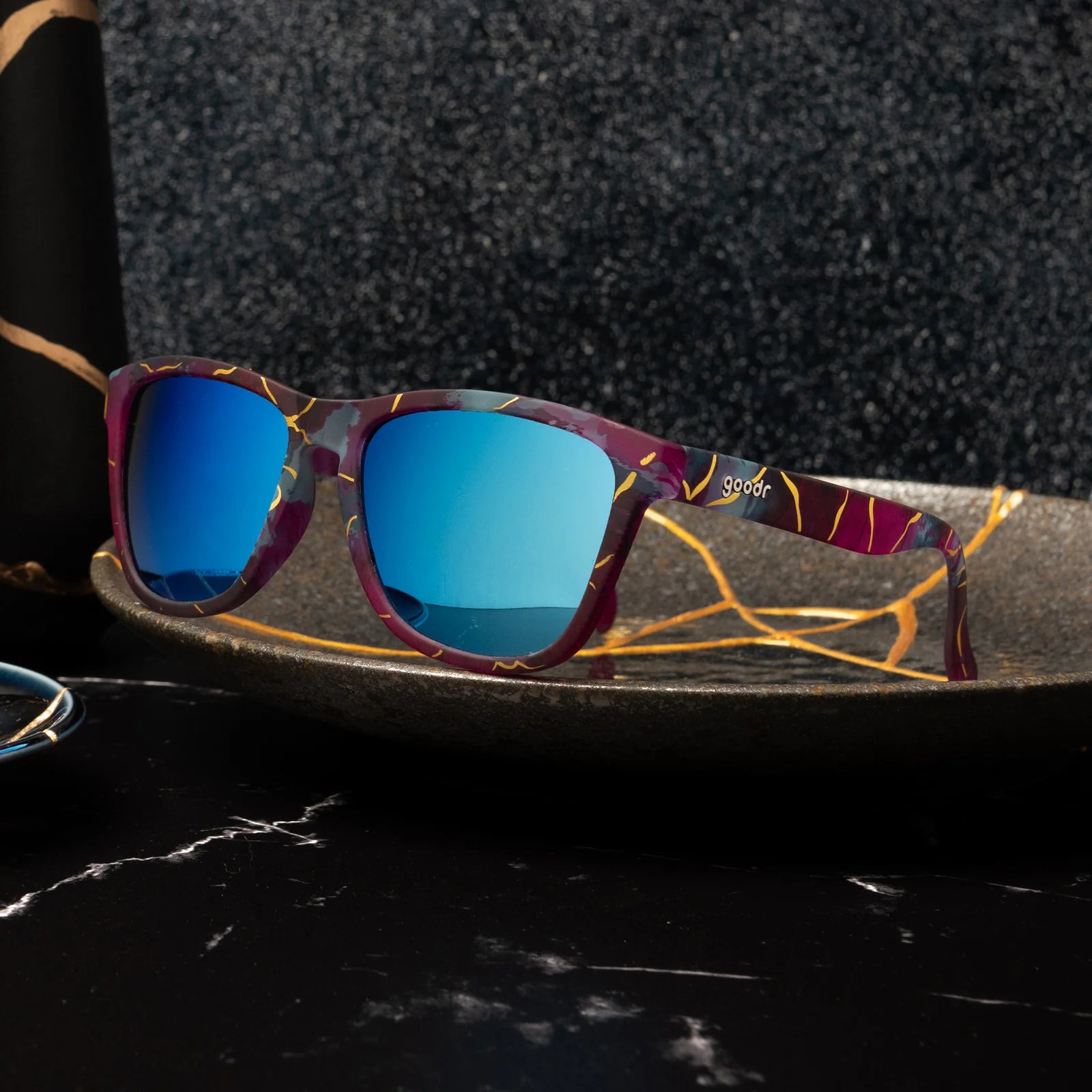 Multi-coloured sunglasses with a prominent gold line pattern. Pinks, purples, and blues on these square-shaped frames.
