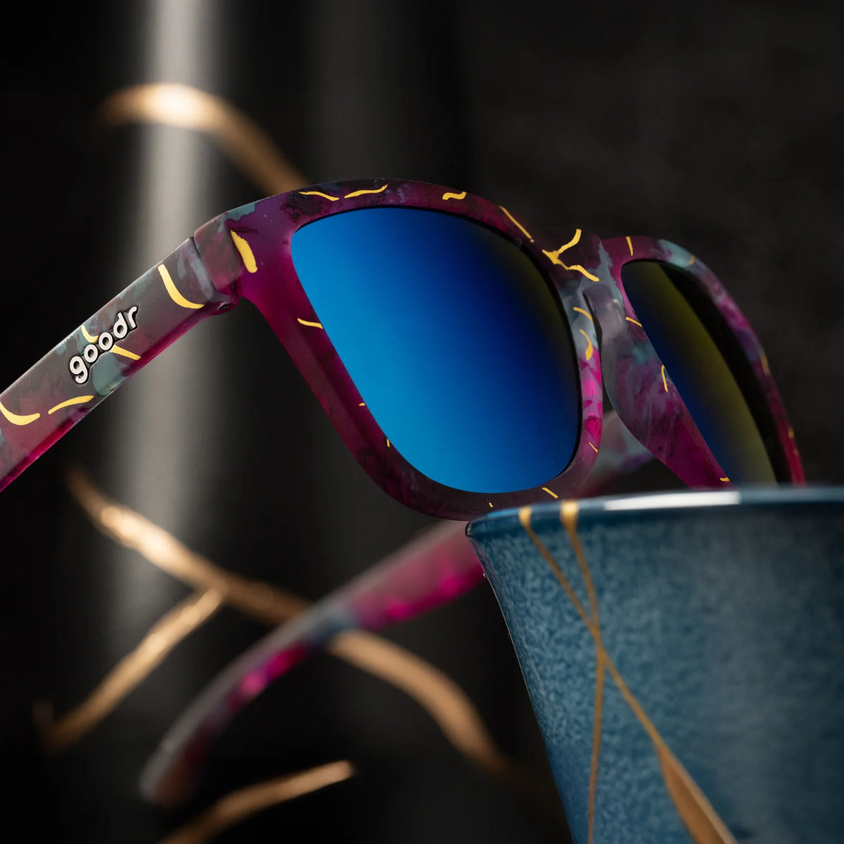 Multi-coloured sunglasses with a prominent gold line pattern. Pinks, purples, and blues on these square-shaped frames.
